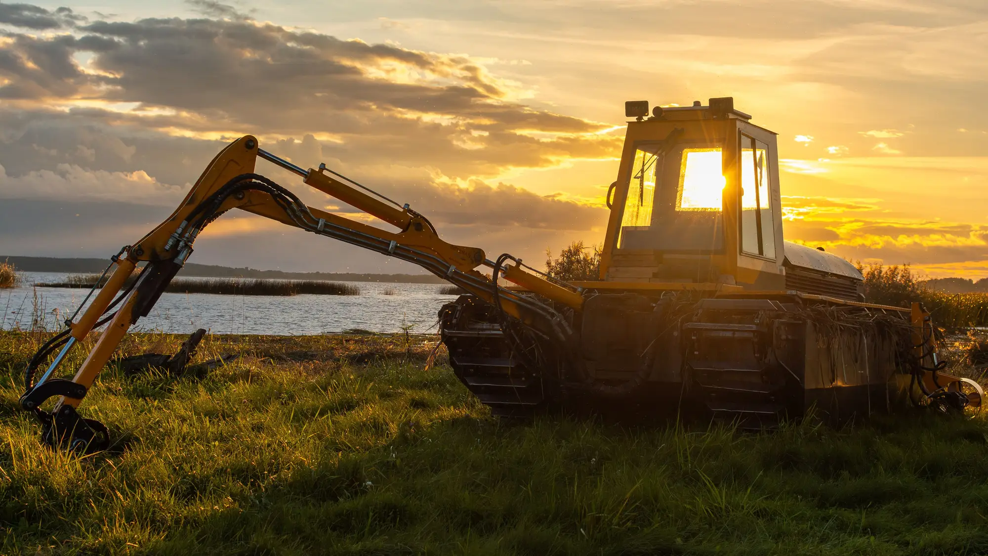 A tracked excavator standing on rough grass at the edge of a lake, low sun behind its cab